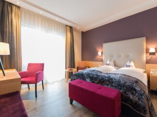 Romantisches Zimmer mit Doppelbett, violetten Akzenten, Schreibtisch, Stühlen, großen Fenstern und sanfter Beleuchtung für ein Candlelight Dinner für zwei Personen.