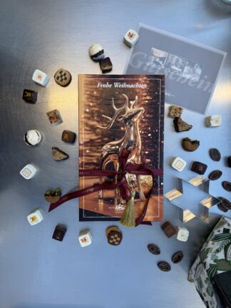 Eine festliche Karte sitzt bei Adventskalender-Pralinenmischung auf einer silbernen Unterlage mit einer Geschenkkarte und Schleife.