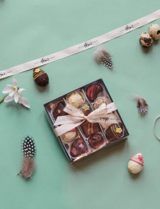 Eine Schachtel mit verschiedenen Pralinen mit einer Schleife, umgeben von Federn, Blumen und verzierten Eiern auf einem hellgrünen Hintergrund.