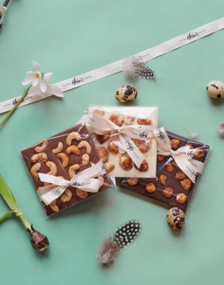 Drei Schokoladentafeln mit Nüssen, eingewickelt in ein Band, umgeben von Federn, Blumen und Wachteleiern auf einem mintgrünen Hintergrund.