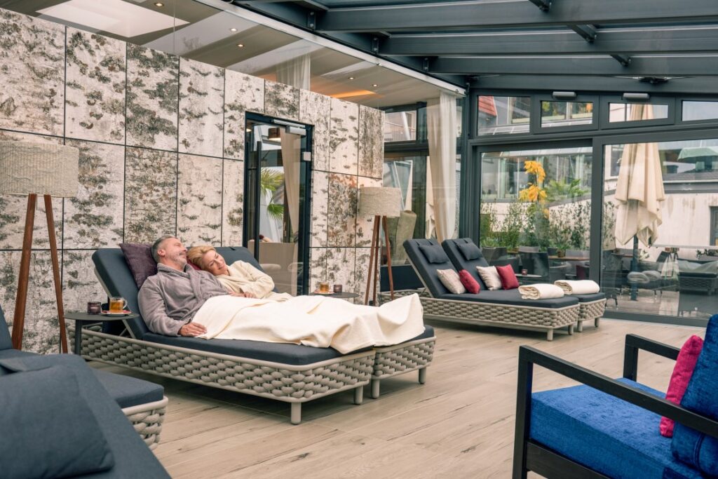 Ein Paar entspannt auf einem Liegestuhl unter Decken in einem modernen Spa-Raum mit großen Fenstern und gemütlichen Sitzgelegenheiten.