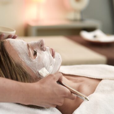 Eine Person liegt auf einer Spa-Liege, während ein Spa-Profi ihr mit einem Pinsel eine Gesichtsmaske aufträgt.