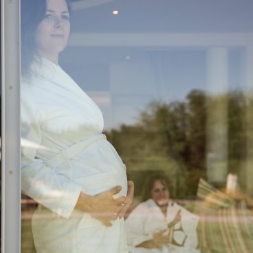 Eine schwangere Frau im weißen Bademantel steht an einem Fenster, im Hintergrund entspannt sich eine andere Frau draußen auf einem Sofa.