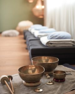Messingklangschalen und Schlägel auf einem Handtuch in einem ruhigen Raum mit gefalteten Decken und Kissen auf Bänken im Hintergrund.