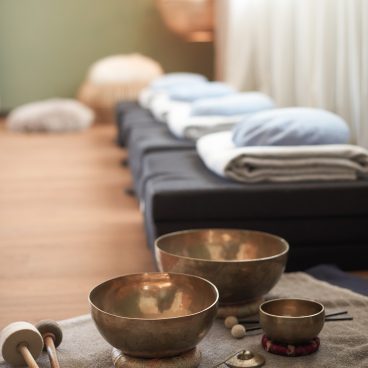 Messingklangschalen und Schlägel auf einem Handtuch in einem ruhigen Raum mit gefalteten Decken und Kissen auf Bänken im Hintergrund.