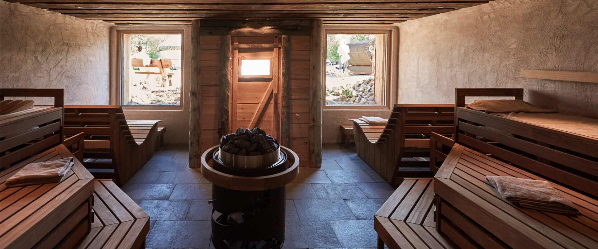 Ein rustikaler Saunaraum aus Holz mit Bänken, einem Zentralofen mit Steinen und Sonnenlicht, das durch drei Fenster strömt.