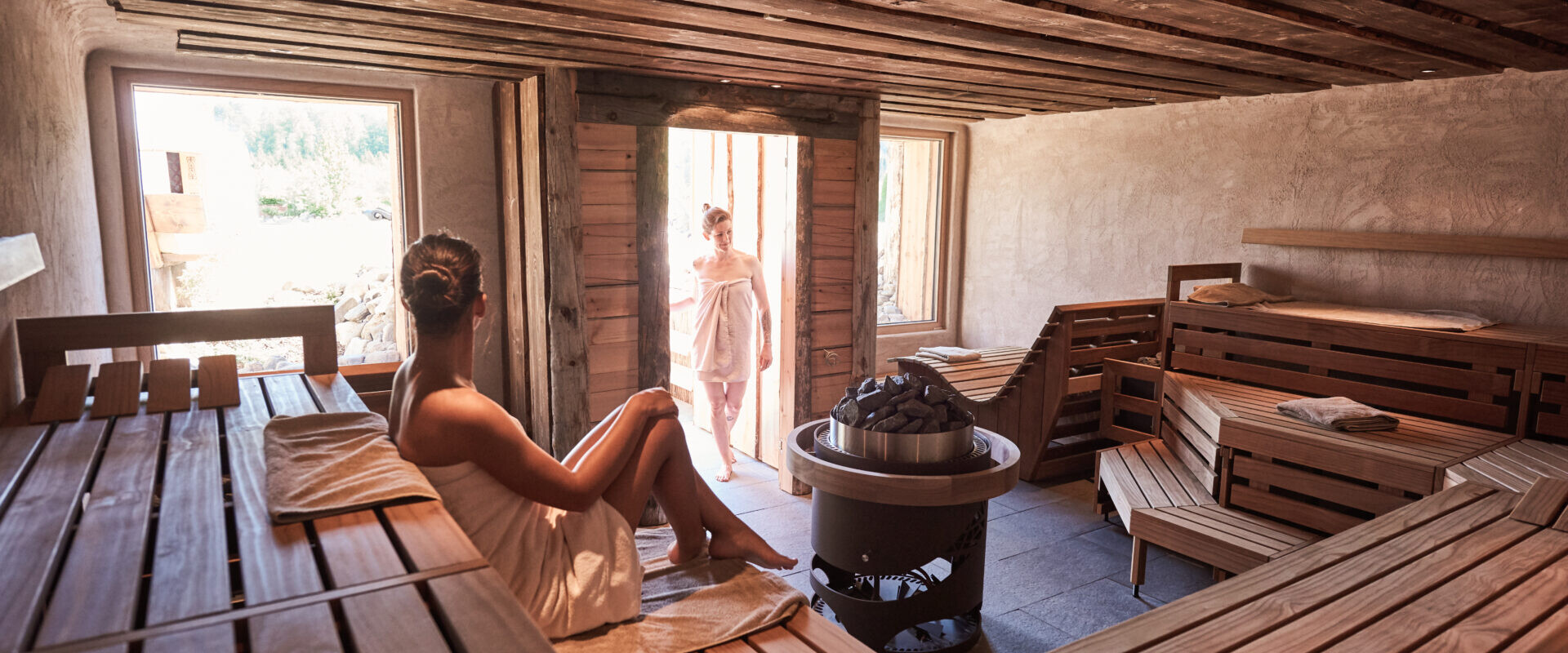 Eine Frau entspannt sich in einer Holzsauna, sie sitzt auf einer Bank, während eine andere, in ein Handtuch gehüllte Frau durch die Tür kommt.
