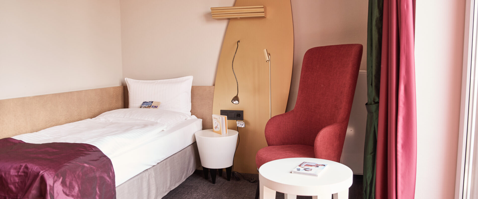 Ein gemütliches Hotelzimmer mit einem Einzelbett, einem roten Sessel, einem kleinen weißen Tisch und einer Wanddekoration in Surfbrettform über dem Nachttisch.