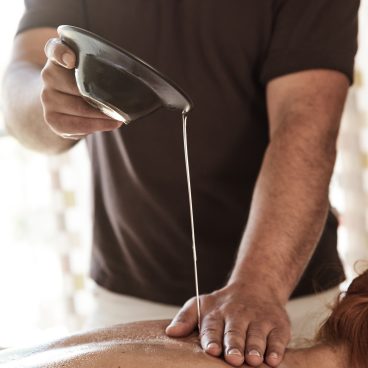 Eine Person gießt während einer Massage Öl aus einer Schüssel auf den Rücken einer anderen Person.
