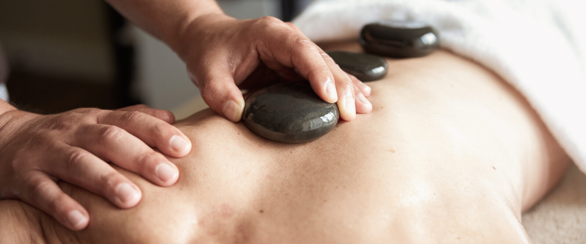 Eine Person erhält eine Hot-Stone-Massage, bei der glatte Steine auf den nackten Rücken gelegt werden und ein Handtuch den Unterkörper bedeckt.