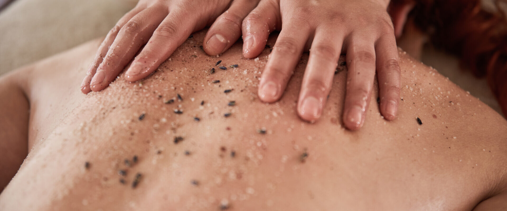 Hände massieren den Rücken einer Person, während auf der Haut ein Peeling mit Salz und getrockneten Kräutern aufgetragen wird.