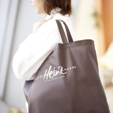 Eine Frau im weißen Bademantel trägt eine große dunkle Einkaufstasche mit der Aufschrift „Hotel Heinz“ und steht im Haus an einem Fenster.