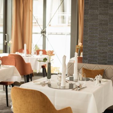 Elegantes Restaurant mit gedeckten Tischen, weißen Tischdecken, orangefarbenen Stühlen und einzelnen Rosen als Tafelaufsatz.