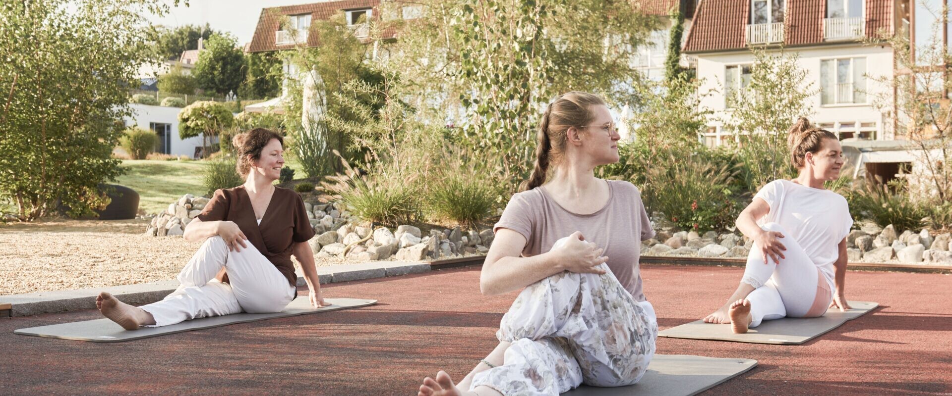 Drei Frauen sitzen an einem sonnigen Tag im Freien auf Yogamatten und machen gemeinsam eine sitzende Drehpose.