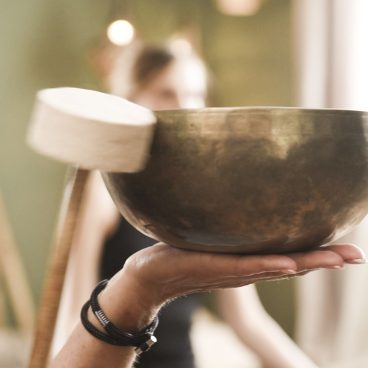 Eine Person hält eine große Klangschale auf der Handfläche, in der anderen Hand einen Schlägel; unscharfer Hintergrund.