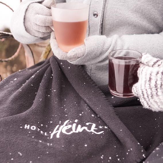 Zwei Personen in Winterhandschuhen halten warme Getränke in der Hand, während sie unter einer schwarzen, mit Schnee bestäubten Decke des Hotels Heinz sitzen.