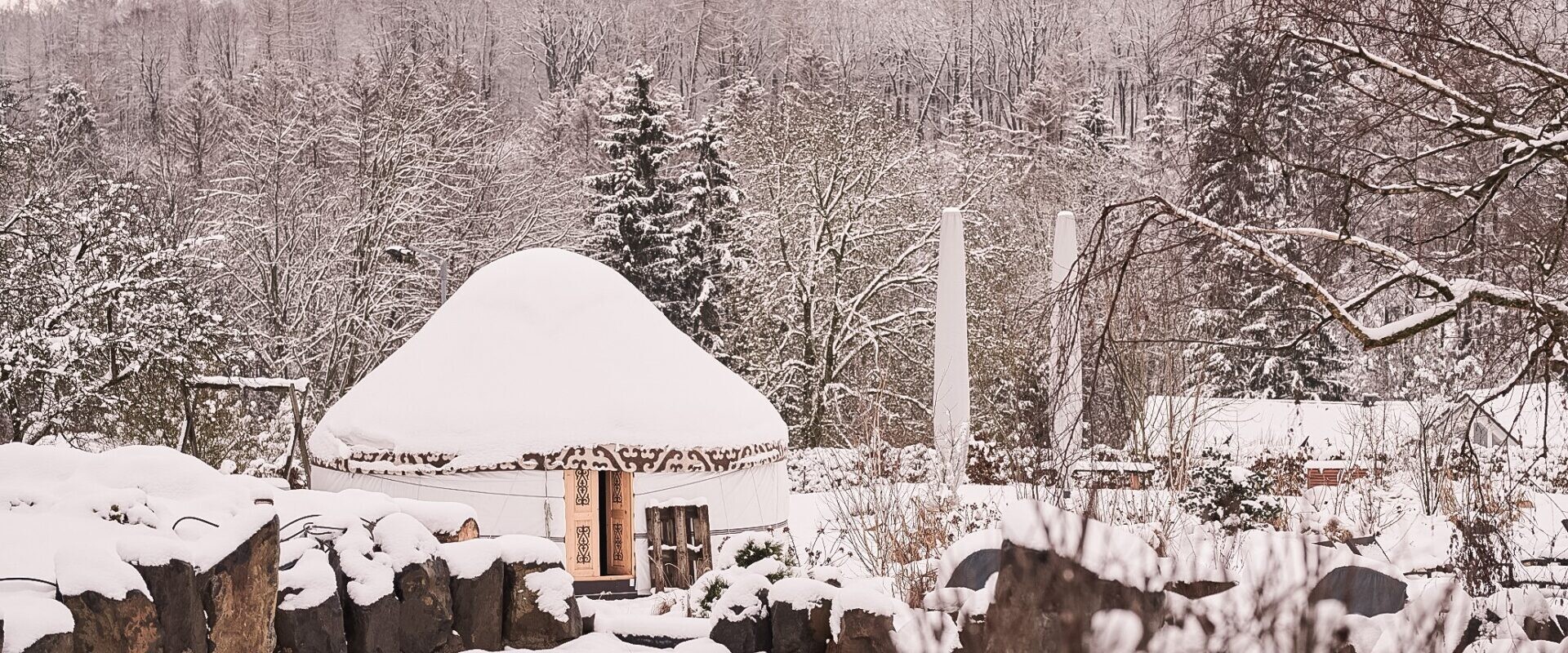 Ein rundes Haus mit Strohdach steht in einer verschneiten Landschaft, umgeben von Bäumen und Felsen, mit einem Wald im Hintergrund.