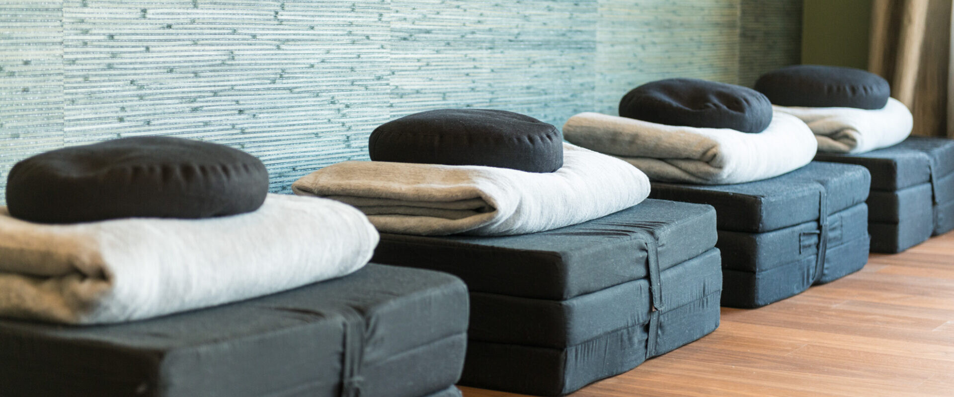 Vier ordentlich gestapelte schwarze Meditationskissen, Matten und gefaltete graue Decken auf einem Holzboden vor einer strukturierten Wand.