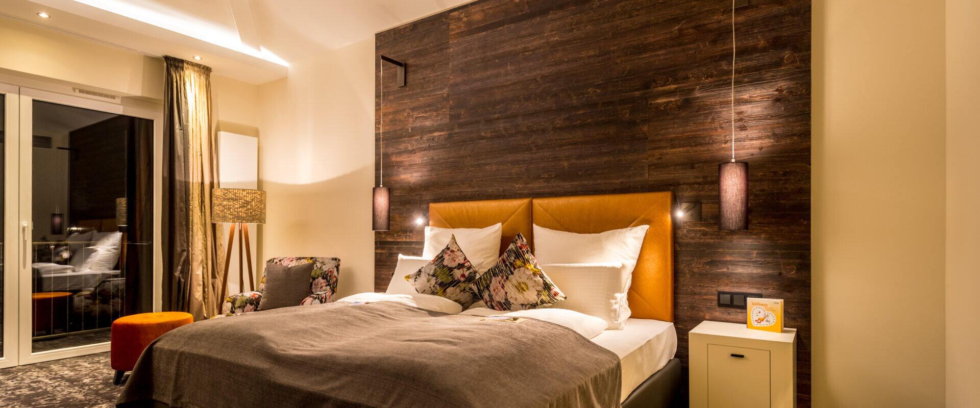 Ein gemütliches, modernes Schlafzimmer mit einem Doppelbett, Kissen mit Blumenmuster, warmer Beleuchtung und einer Akzentwand aus dunklem Holz hinter dem Kopfteil.