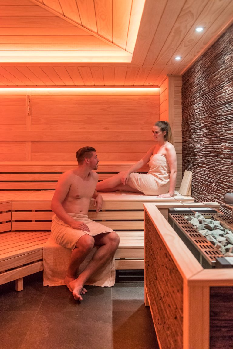 Ein Mann und eine Frau in Handtüchern entspannen und unterhalten sich in einer modernen Holzsauna mit warmer Beleuchtung und Steinwänden.