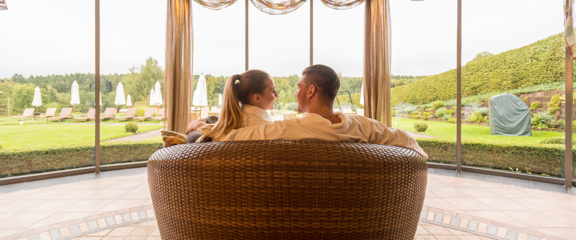 Ein Paar sitzt zusammen auf einem runden Korbsofa vor großen Fenstern mit malerischem Blick auf den Garten.