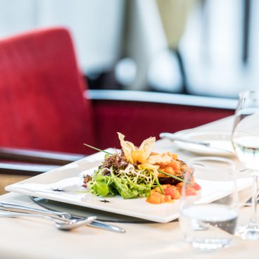 Ein Gourmetsalat mit Blattgemüse, Gemüse und Garnitur auf einem gedeckten Tisch mit einem Glas Wasser und Wein in einem hellen Restaurant.