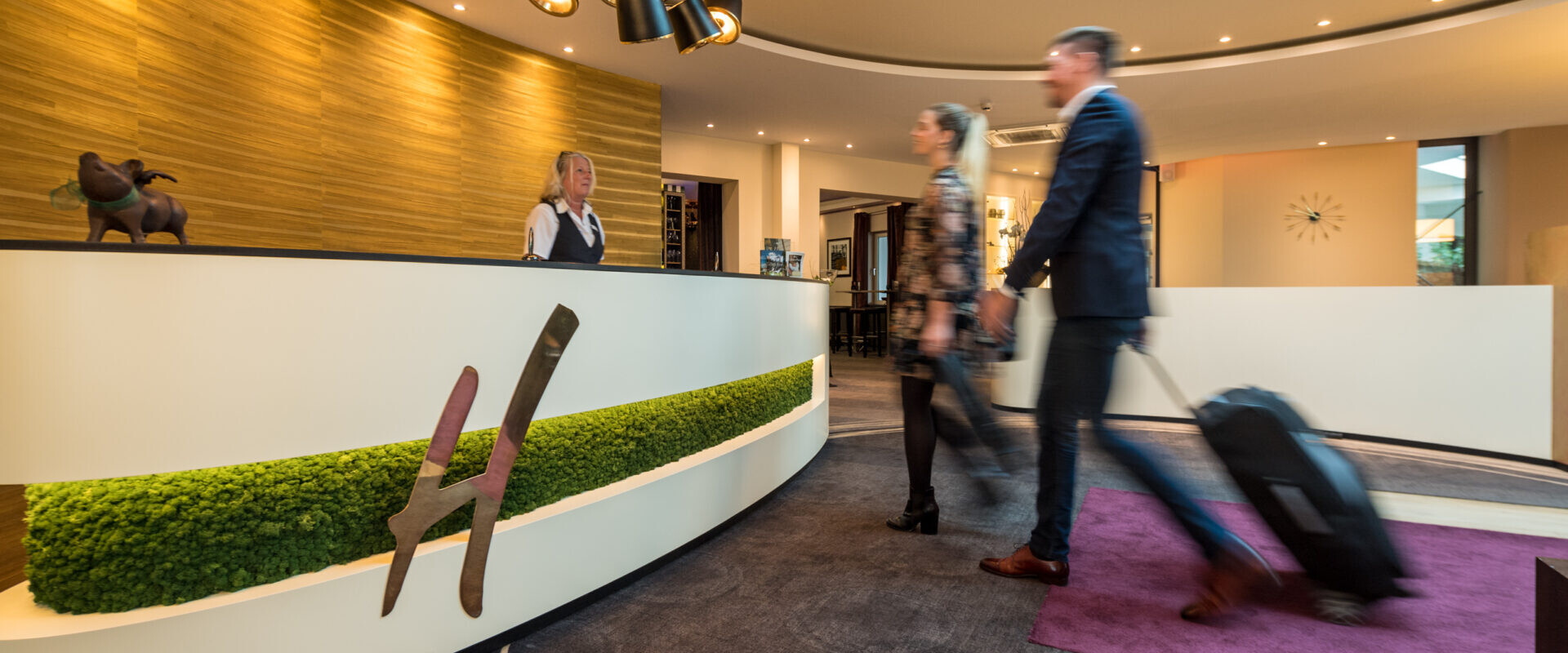 Zwei Personen mit Gepäck nähern sich einer modernen Hotelrezeption, wo ein Mitarbeiter bereitsteht, um ihnen zu helfen.