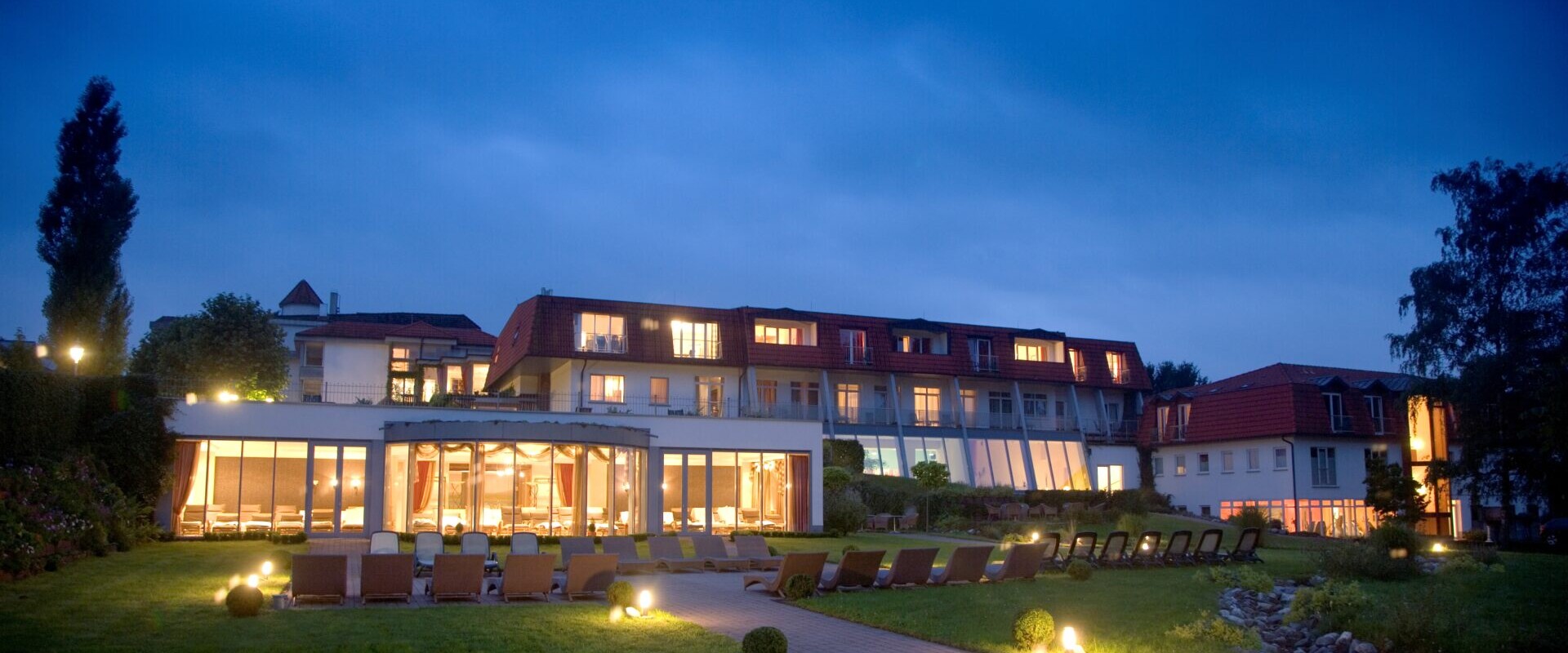 Modernes Hotelgebäude in der Abenddämmerung mit leuchtenden Lichtern, großen Fenstern und Liegestühlen auf einer Rasenfläche unter einem bewölkten Abendhimmel.