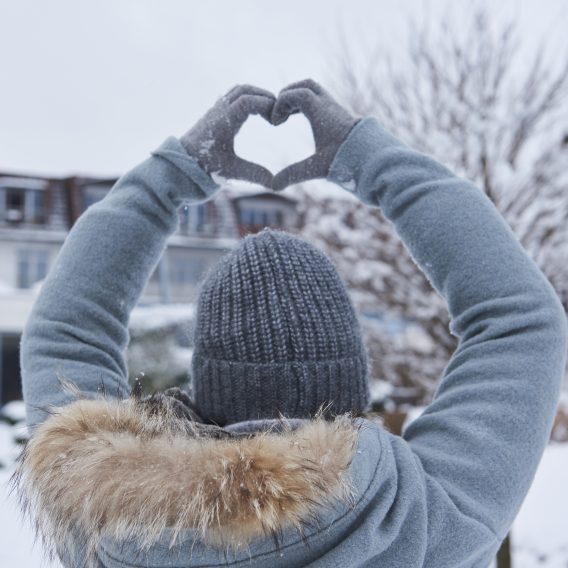 Eine Person in Wintermantel und Mütze hält behandschuhte Hände über dem Kopf und formt in einer verschneiten Umgebung im Freien ein Herz.
