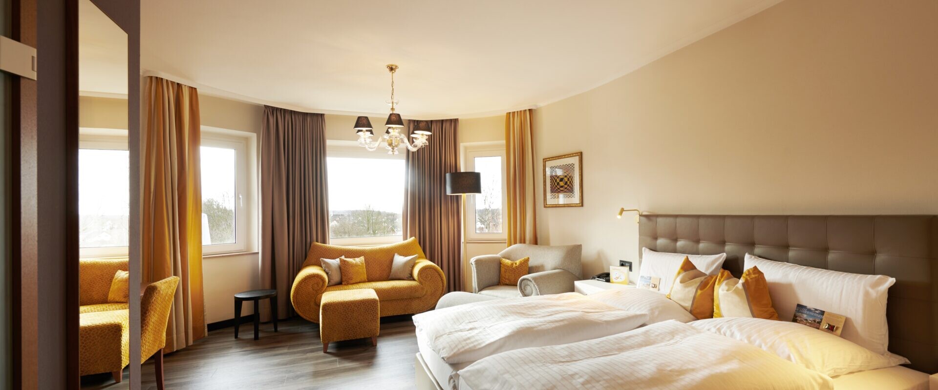 Geräumiges Hotelzimmer mit Doppelbett, gelbem Sofa, Sessel, Kronleuchter, großen Fenstern und warmer Beleuchtung.