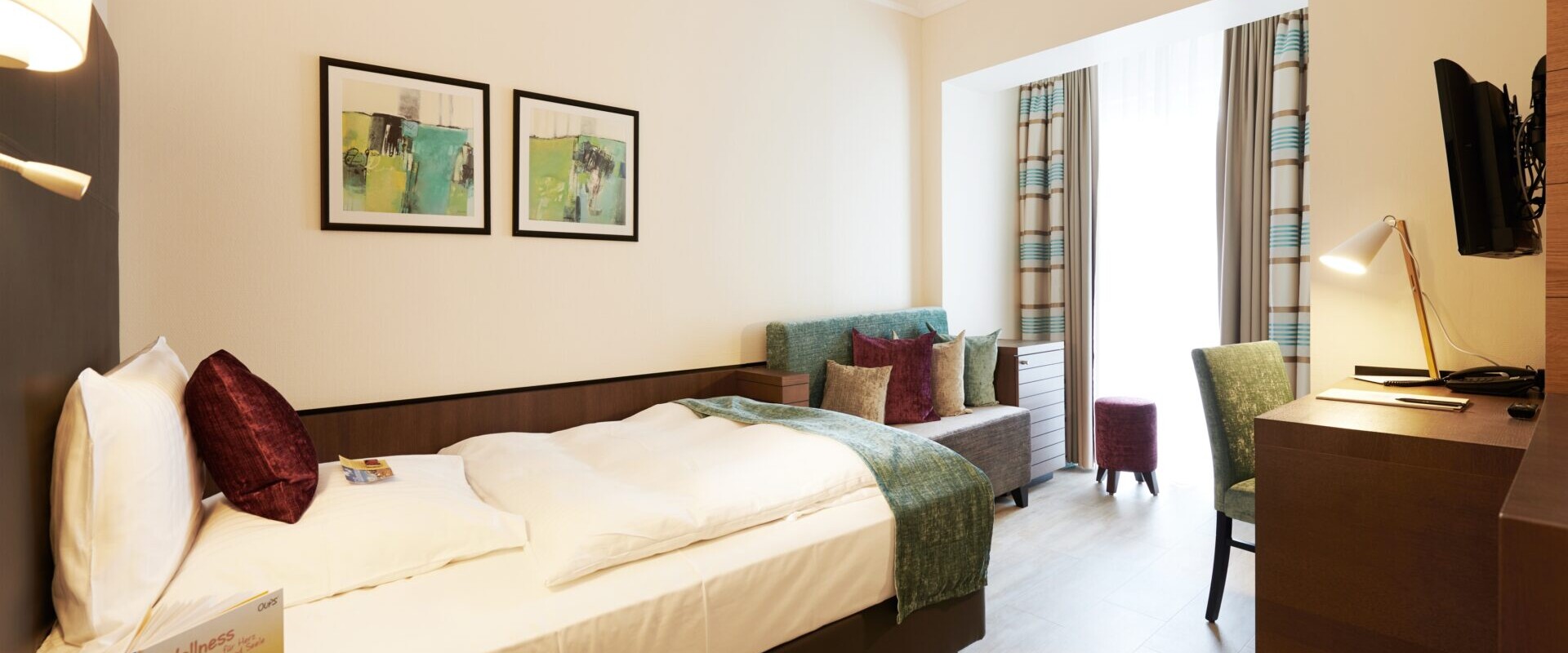 Ein helles Hotelzimmer mit Doppelbett, Sofa, Schreibtisch, Fernseher und großem Fenster mit Vorhängen, die Tageslicht hereinlassen.