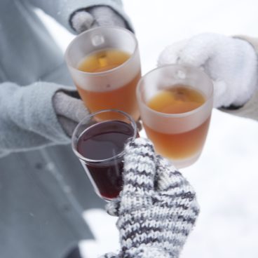 Drei Personen mit Winterhandschuhen stoßen im Freien in einer verschneiten Umgebung mit warmen Getränken an.