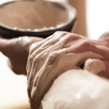 Eine Nahaufnahme von Händen, die eine Maske oder Substanz aus weißem Ton auf einen Arm auftragen, mit einer Keramikschale im Hintergrund.