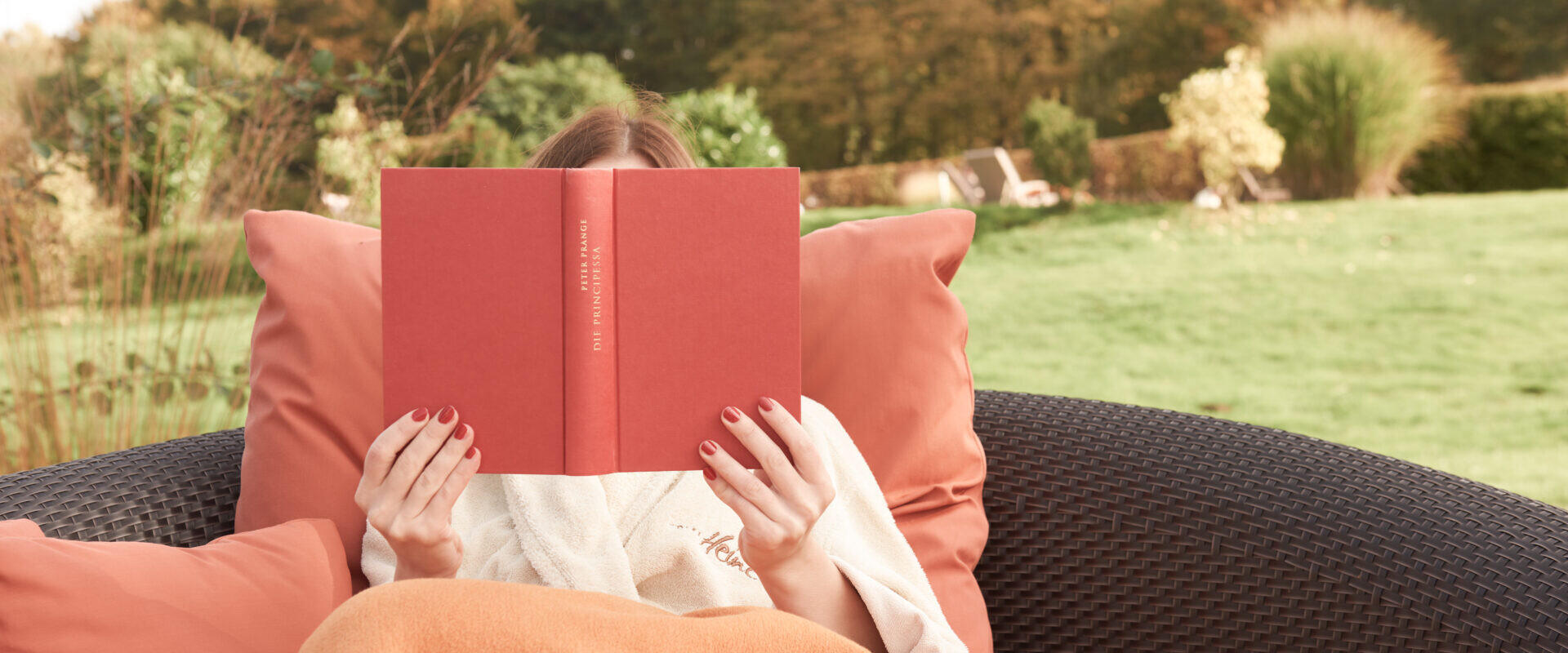 Person, die im Freien auf einem Korbstuhl sitzt, in eine orangefarbene Decke gehüllt, ein rotes Buch liest, im Hintergrund sind Herbstbäume zu sehen.