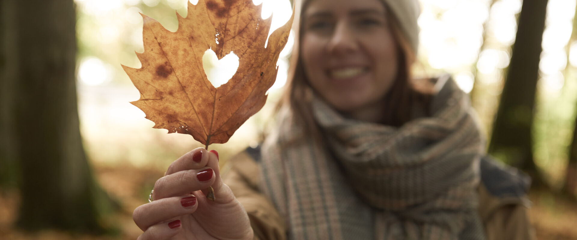 Herbst-Urlaub im Westerwald. Eine Frau hält ein braunes Blatt mit einem herzförmigen Loch und lächelt in einem Wald, während sie einen Schal und eine Mütze trägt.