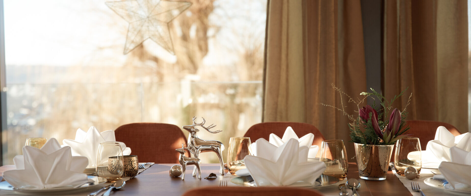 Festlicher Esstisch mit gefalteten weißen Servietten, Golddekor, einer Rentierfigur und einer Sternendekoration im Fenster.