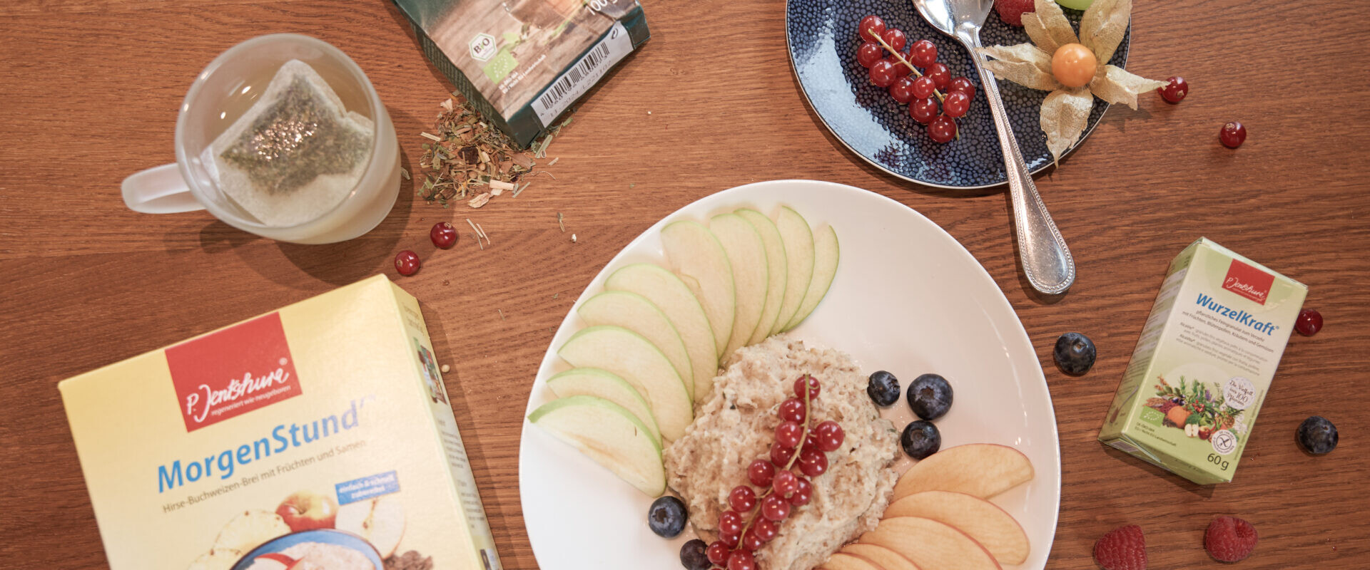 Eine Frühstücksszene mit einem Teller Müsli, Apfelscheiben, Beeren, Tee, einer Teebox und einer Packung Getreide auf einem Holztisch.