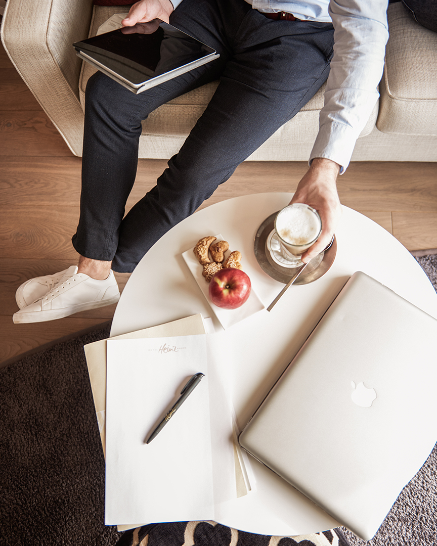 Eine Person sitzt auf einem Sofa und hält ein Tablet in der Hand. Auf einem runden weißen Tisch stehen ein Laptop, ein Notizblock, ein Stift, ein Apfel, Kekse und ein Getränk.