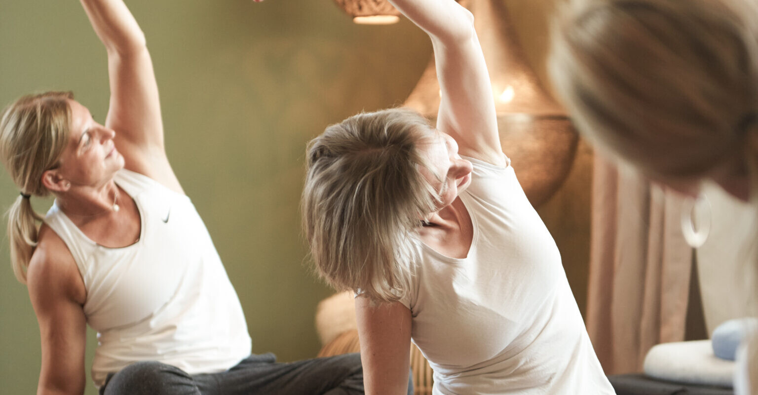 Drei Frauen sitzen im Innenbereich auf Yogamatten und strecken während einer Yogastunde ihre Arme über den Kopf, in der Nähe gibt es warmes Licht und dekorative Lampen.