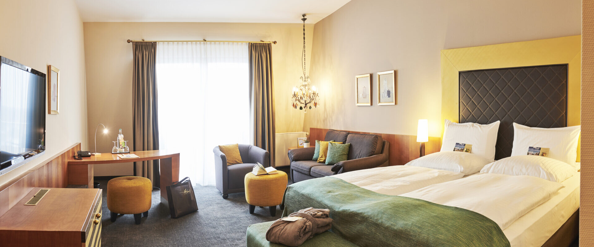 Ein helles Hotelzimmer mit einem großen Bett, einem Sofa, einem Schreibtisch, einem Fernseher und einem hohen Fenster mit Vorhängen, die natürliches Licht hereinlassen.