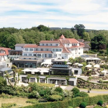 Ein großes, elegantes Hotel mit Gärten und Außenterrassen, an einem sonnigen Tag von Bäumen und Grün umgeben.