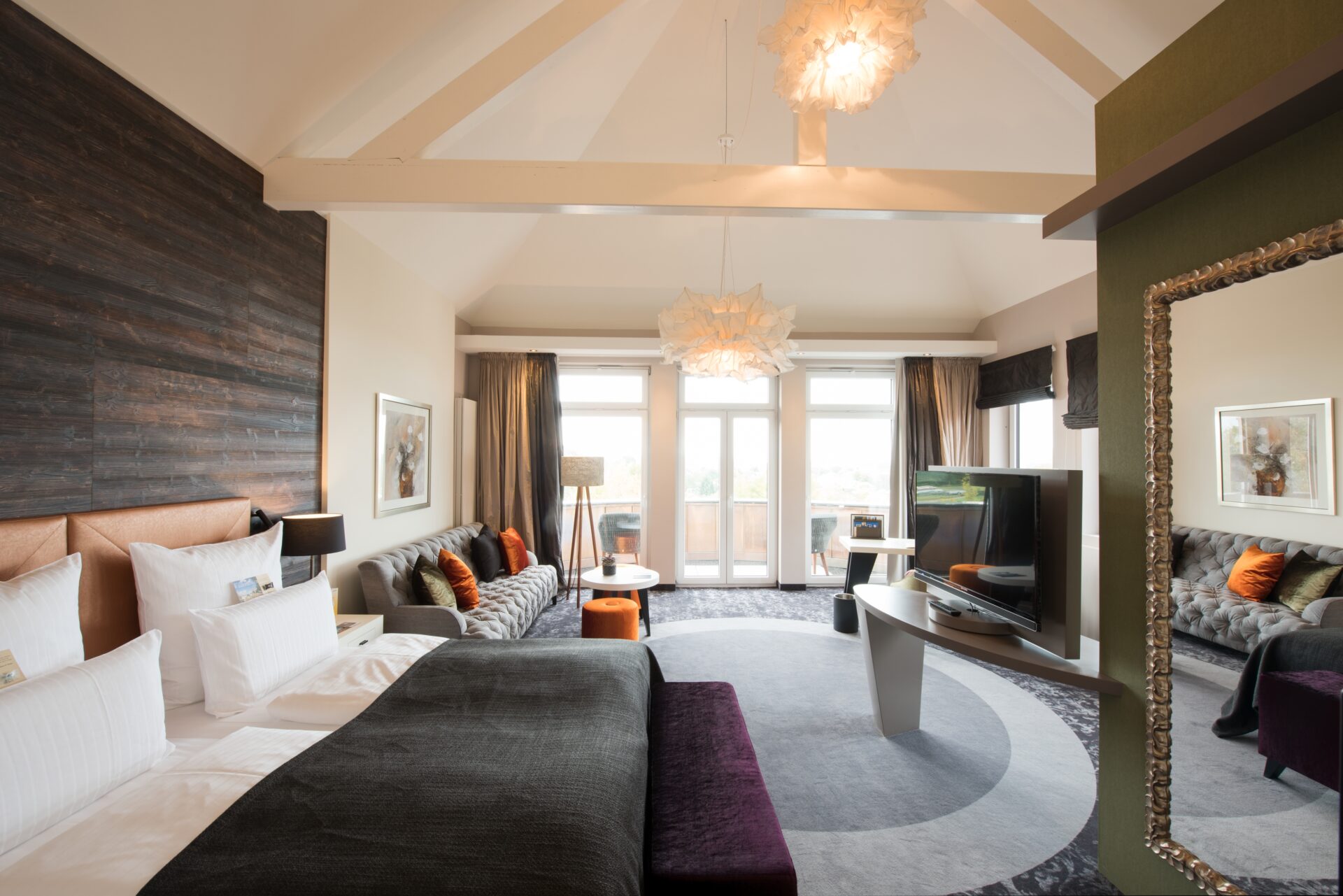 Geräumiges, modernes Hotelzimmer mit großem Bett, Sofa, Fernseher, Schreibtisch und raumhohen Fenstern, die viel Tageslicht hereinlassen.