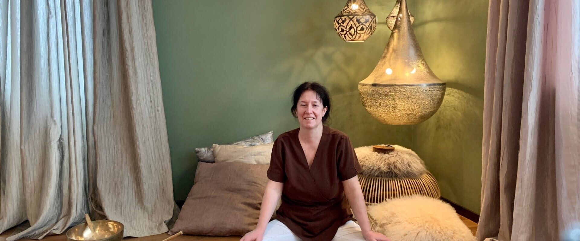 Eine Frau sitzt im Schneidersitz auf einem Holzboden in einem gemütlichen Raum mit Kissen, Klangschalen und hängenden Pendelleuchten.