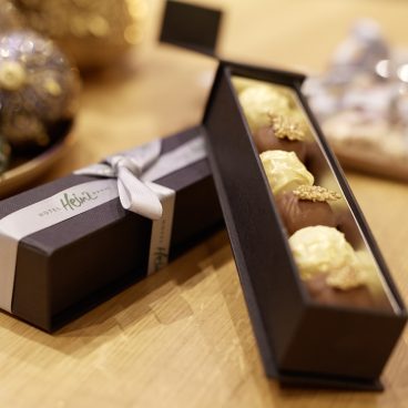 Auf einem Holztisch steht eine schwarze Geschenkbox mit verschiedenen Pralinen, offen und mit einer silbernen Schleife verschnürt.