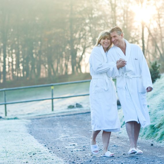 Ein älteres Paar in weißen Gewändern geht Arm in Arm auf einem frostigen Pfad im Freien und lächelt in der Morgensonne.