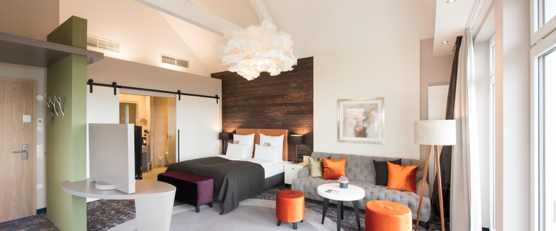 Modernes Hotelzimmer mit Doppelbett, Sofa, Fernseher, Schreibtisch und großen Fenstern, die Tageslicht hereinlassen, dekoriert mit orangefarbenen Akzenten.