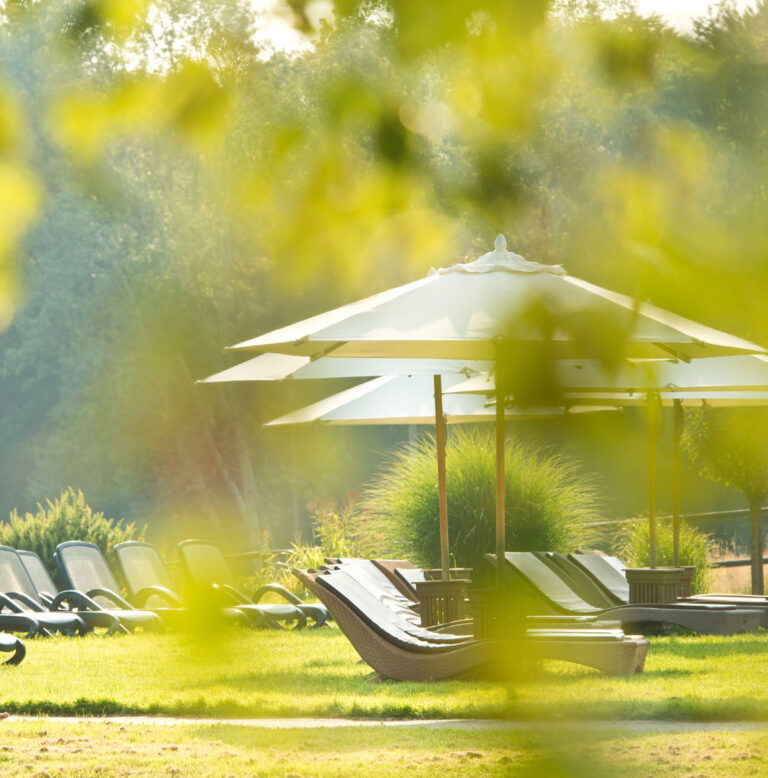 Ein älteres Paar geht zusammen auf einer sonnigen Wiese spazieren, umgeben von Grün, in der Nähe von leeren Liegestühlen und weißen Sonnenschirmen.