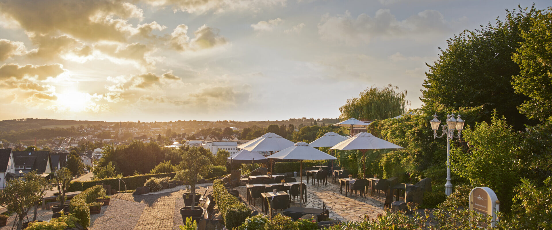 Von der Außenterrasse des Restaurants mit Tischen und Sonnenschirmen hat man bei Sonnenuntergang einen Blick auf eine malerische Landschaft, umgeben von üppigem Grün und einem Kopfsteinpflasterweg.