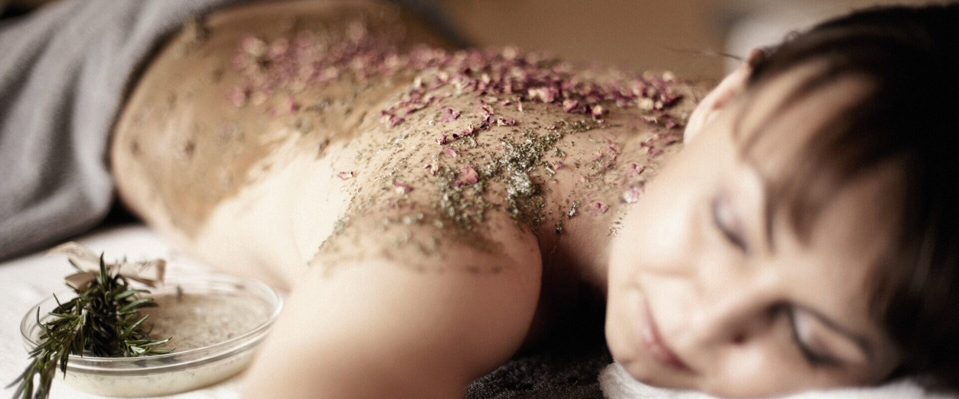 Eine Frau entspannt sich bäuchlings auf einem Massagetisch, während ihr Rücken mit einem Kräuter- und Blütenpeeling behandelt wird.