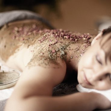 Eine Frau entspannt sich bäuchlings auf einem Massagetisch, während ihr Rücken mit einem Kräuter- und Blütenpeeling behandelt wird.